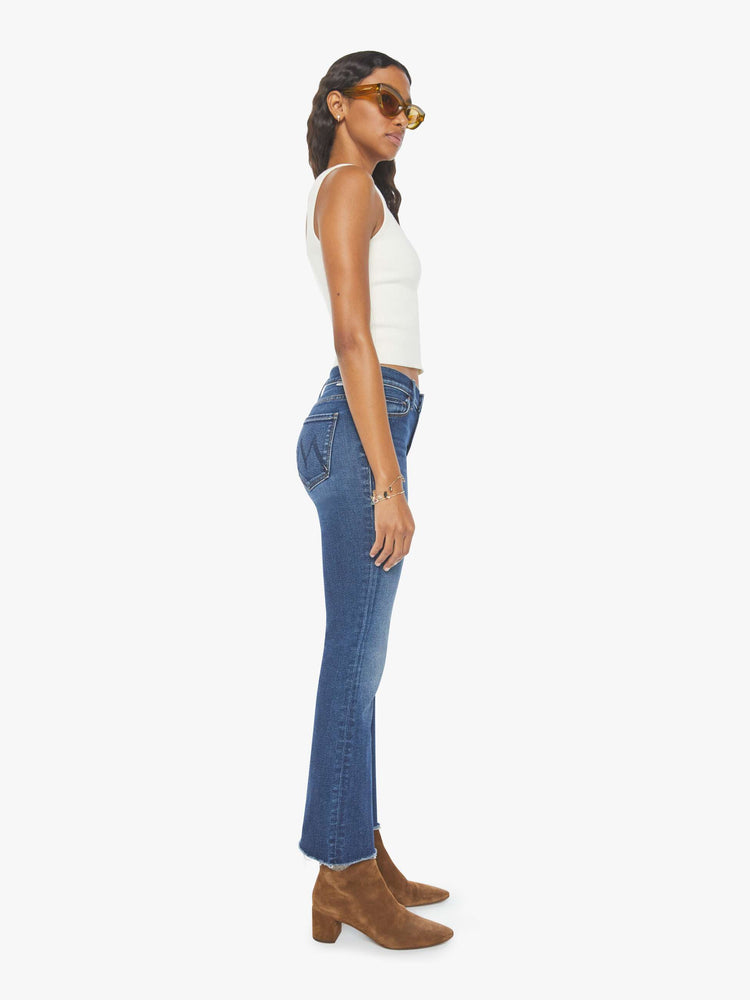 Side view of a woman narrow flare jeans with a mid rise and an ankle-length inseam with a frayed hem in a dark blue wash.