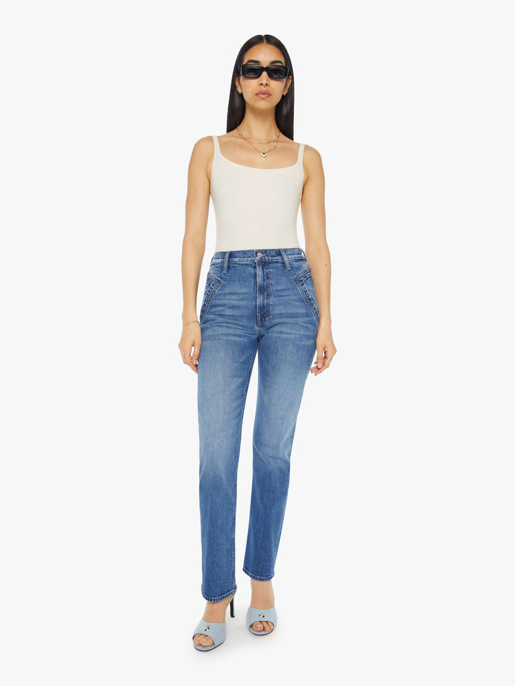 Front view of a woman in a mid blue high rise jean with a narrow straight leg, triangle pockets and a 31-inch inseam with a clean hem.