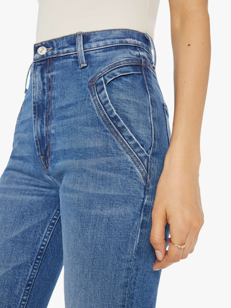 Close up view of a woman in a mid blue high rise jean with a narrow straight leg, triangle pockets and a 31-inch inseam with a clean hem.