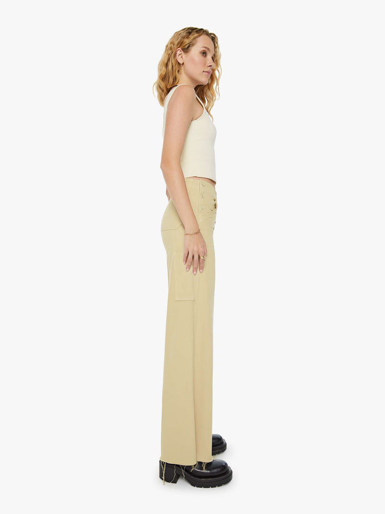 Side view of a woman in super high waisted light khaki wide-leg pants with buttoned details at the hips, utility-inspired patch pockets, a hammer loop and a long 32-inch inseam with a frayed hem. 