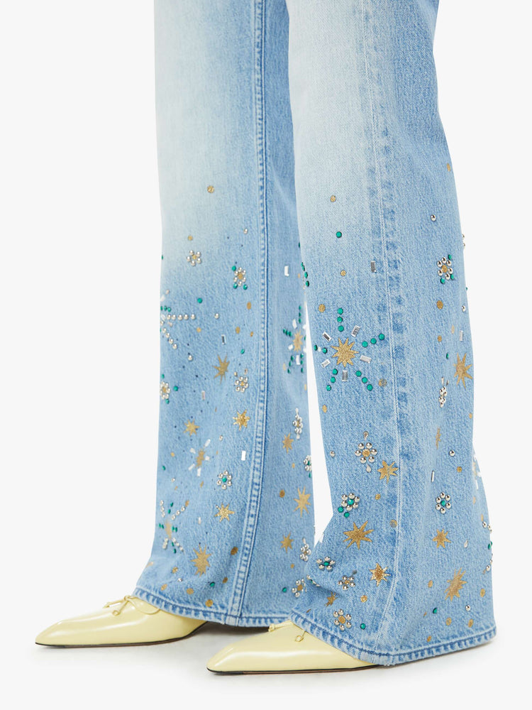 Hem close up view of a woman in a light blue high rise bootcut jean embellished with paint, glitter and studs.