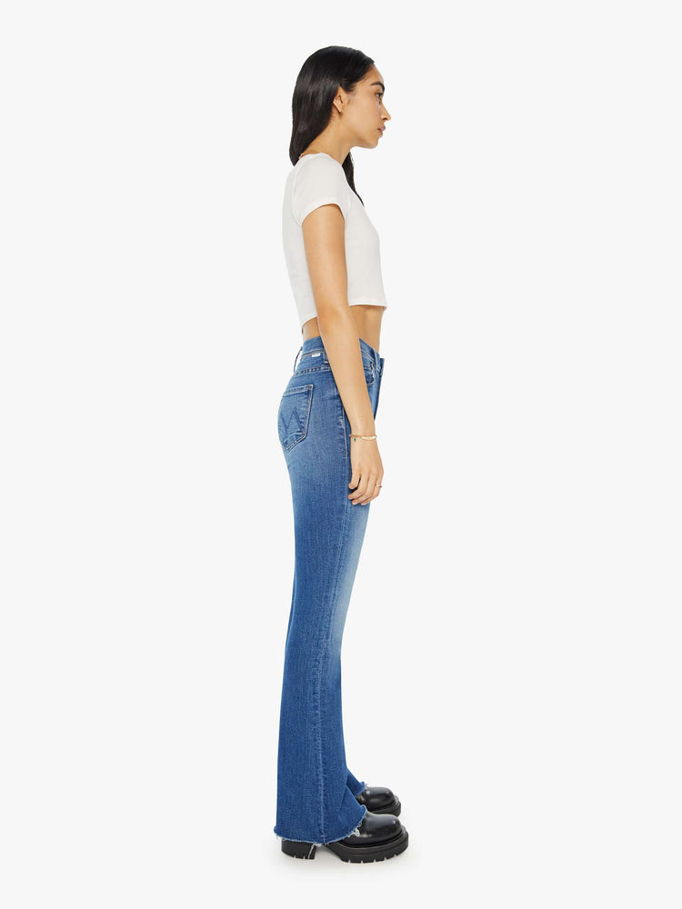 Side view of a woman in mid-blue md-rise jeans with a flared leg, long 32-inch inseam and a frayed hem.