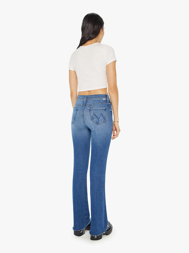 Back view of a woman in mid-blue md-rise jeans with a flared leg, long 32-inch inseam and a frayed hem.