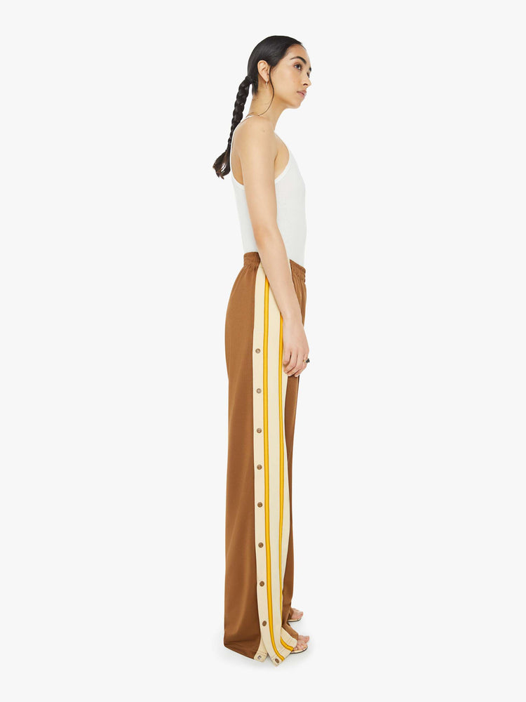 Side view of a woman in track pant features a high rise, wide leg, elastic waistband, slit pockets that zip, a long 34-inch inseam and a seamed detail down the front in light brown.