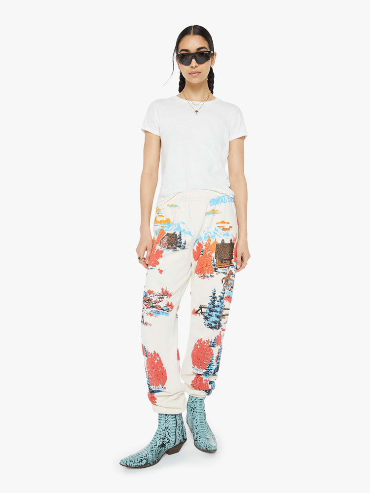 Front view of a woman in sweatpants with a loose straight leg, long inseam and an elastic waist and hems in off white with a tranquil puff-paint graphic of horses, cabins and trees down the legs.