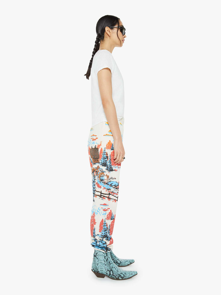 Side view of a woman in sweatpants with a loose straight leg, long inseam and an elastic waist and hems in off white with a tranquil puff-paint graphic of horses, cabins and trees down the legs.