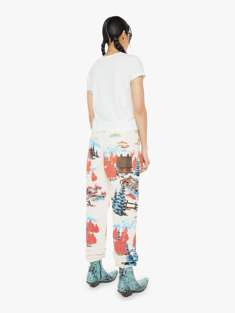 Back view of a woman in sweatpants with a loose straight leg, long inseam and an elastic waist and hems in off white with a tranquil puff-paint graphic of horses, cabins and trees down the legs.