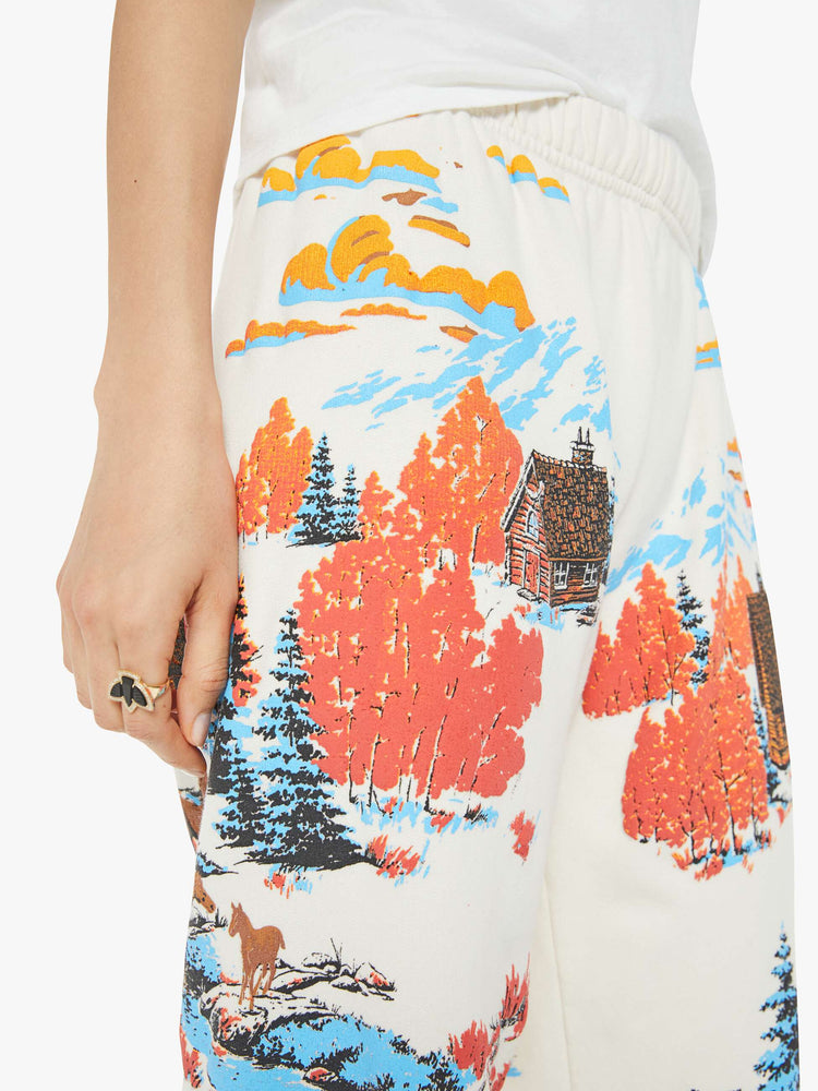 Close up waist view of a woman in sweatpants with a loose straight leg, long inseam and an elastic waist and hems in off white with a tranquil puff-paint graphic of horses, cabins and trees down the legs.