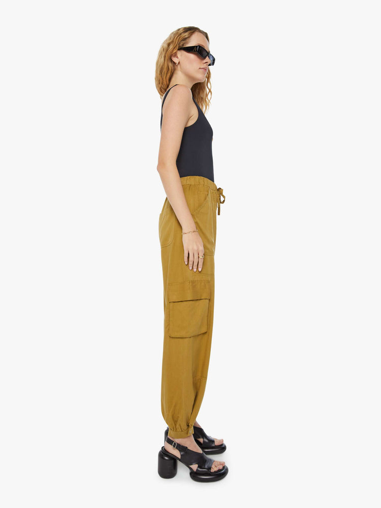 Side view of a woman in a bronze yellow high waisted cargo pant with an elastic drawstring waist, barrel leg, patch pockets and a long inseam with snaps at the hem.