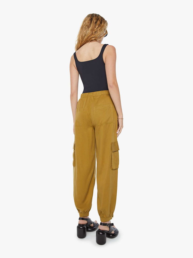 Back view of a woman in a bronze yellow high waisted cargo pant with an elastic drawstring waist, barrel leg, patch pockets and a long inseam with snaps at the hem.