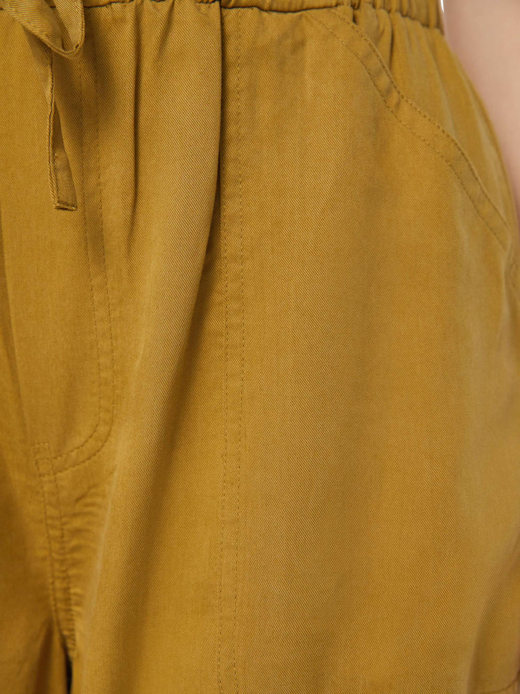 Detailed view of a woman in a bronze yellow high waisted cargo pant with an elastic drawstring waist, barrel leg, patch pockets and a long inseam with snaps at the hem.