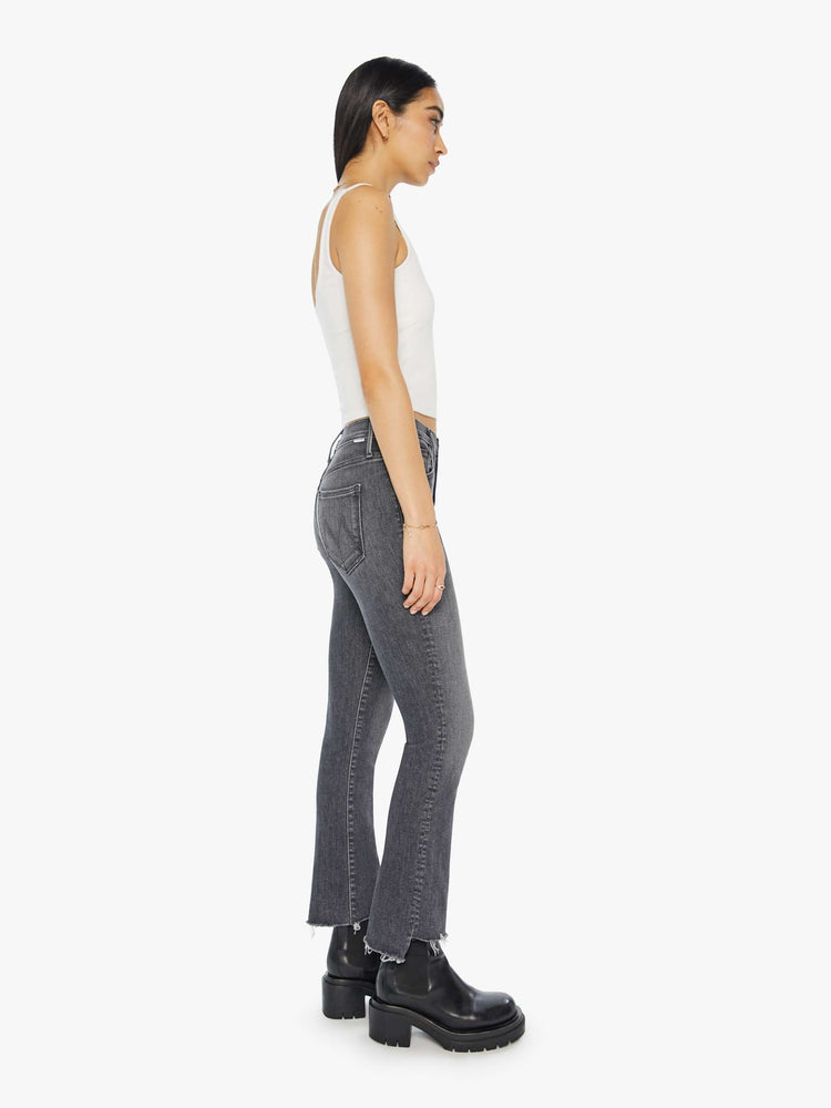 Sideview of a woman in a high waisted bootcut that hits at the ankle with a frayed step hem in a dark grey wash.