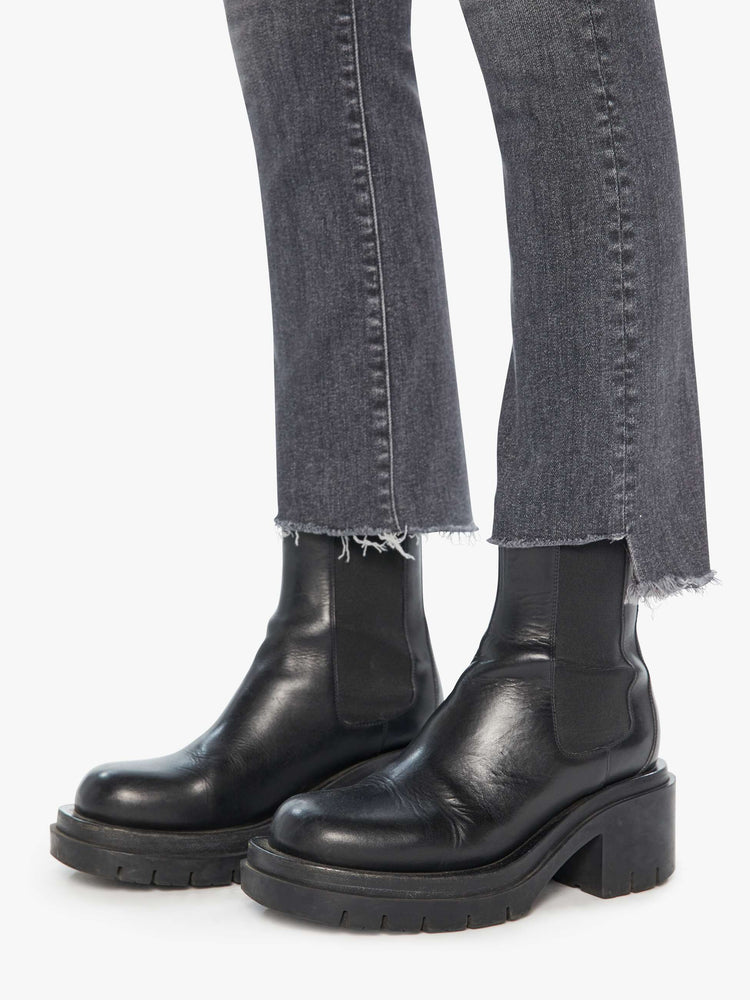 Hem view of a woman in a high waisted bootcut that hits at the ankle with a frayed step hem in a dark grey wash.