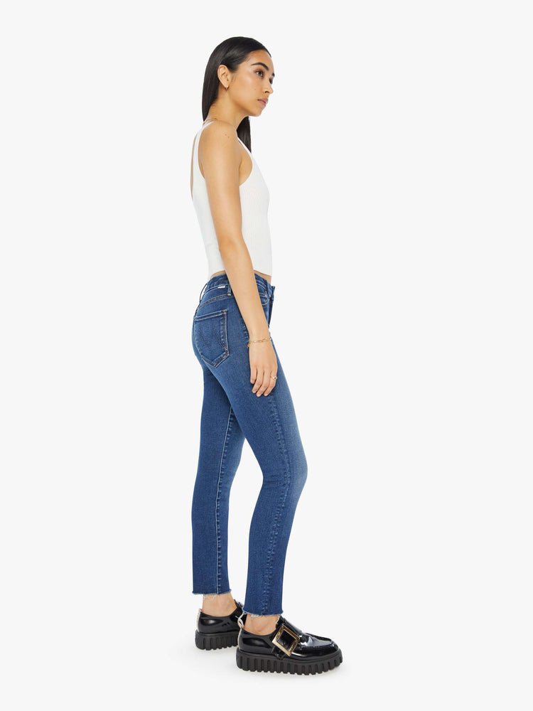 Side view of a woman in a dark blue mid-rise skinny jean with an ankle-length inseam and a frayed hem.