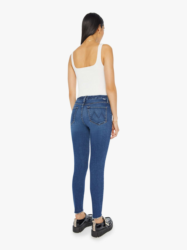 Back view of a woman in a dark blue mid-rise skinny jean with an ankle-length inseam and a frayed hem.