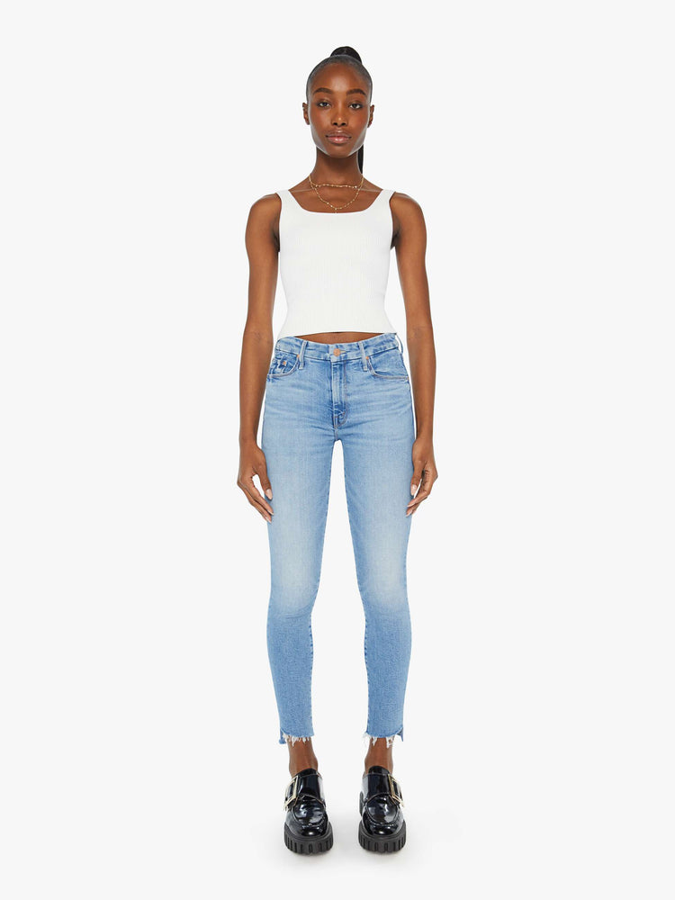 Front view of a woman in a light blue wash high-rise skinny jean has an ankle-length inseam and a frayed step hem.