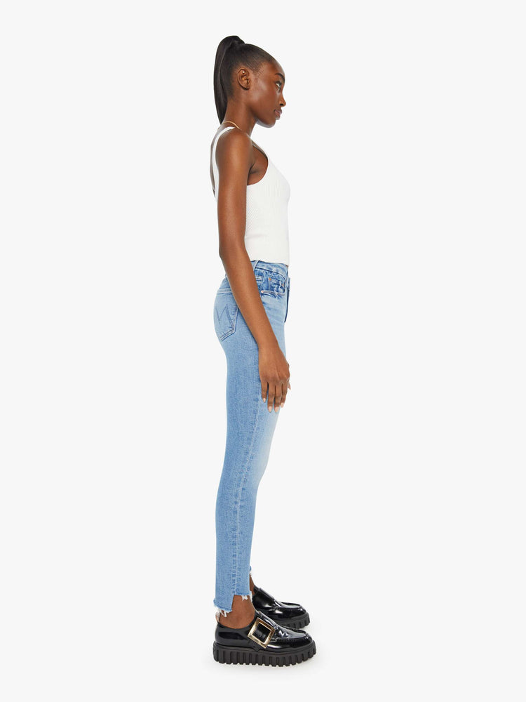 Side view of a woman in a light blue wash high-rise skinny jean has an ankle-length inseam and a frayed step hem.