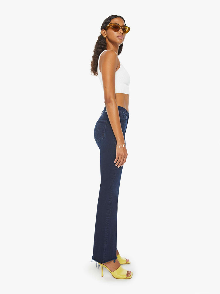 Side view of a woman in a dark blue wash mid-rise flare with a 31-inch inseam and frayed hem.