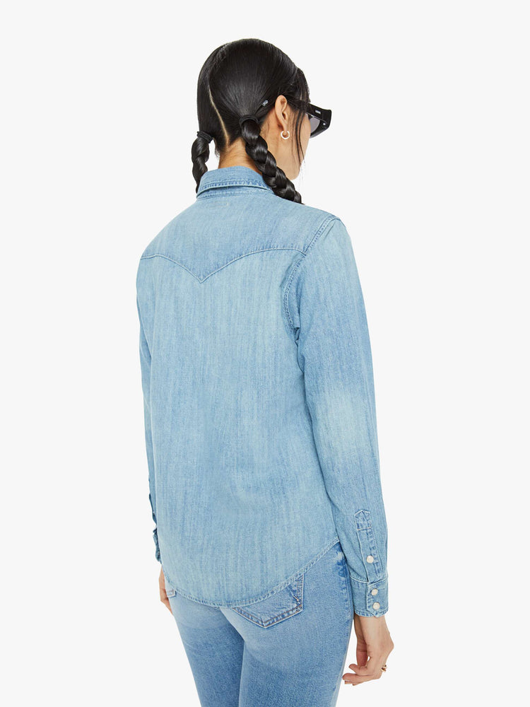 Back view of a woman in denim button-up with front patch pockets and a curved hem in a light blue wash with snap buttons and metal tips on the collar.