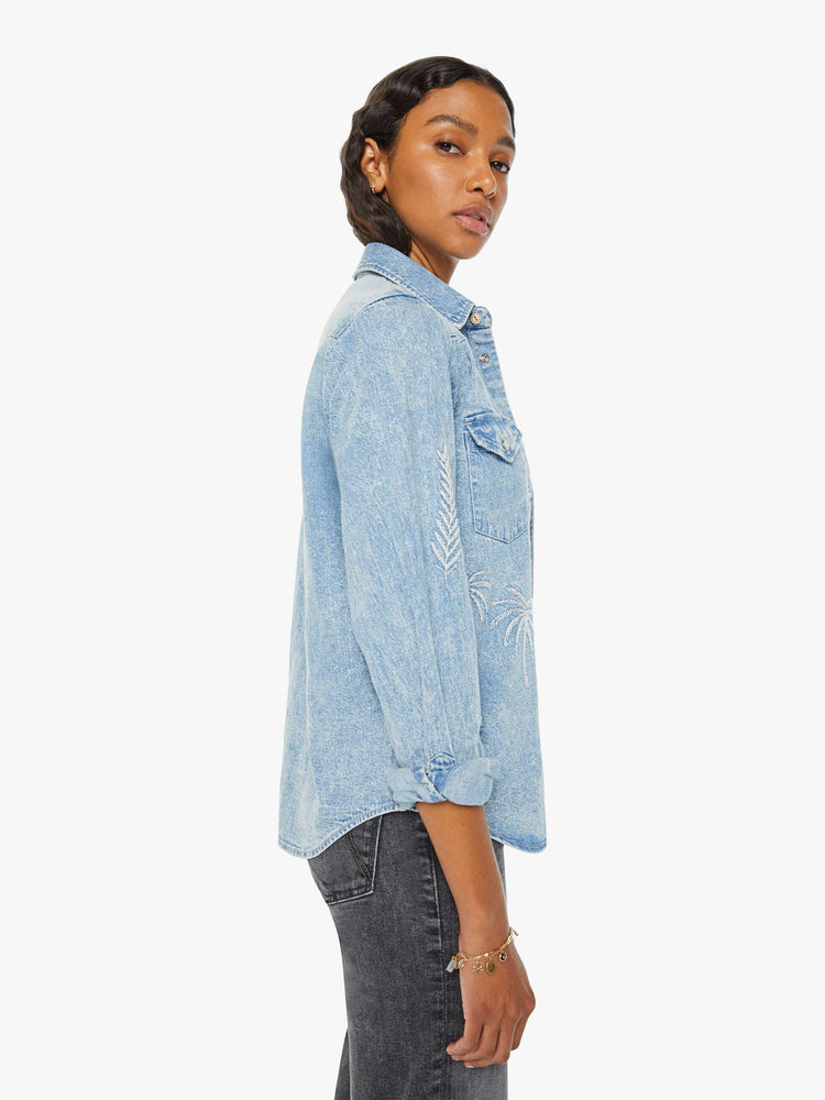 Side view of a woman in a light blue denim button-up with front patch pockets, a curved hem, and embroidered details throughout.