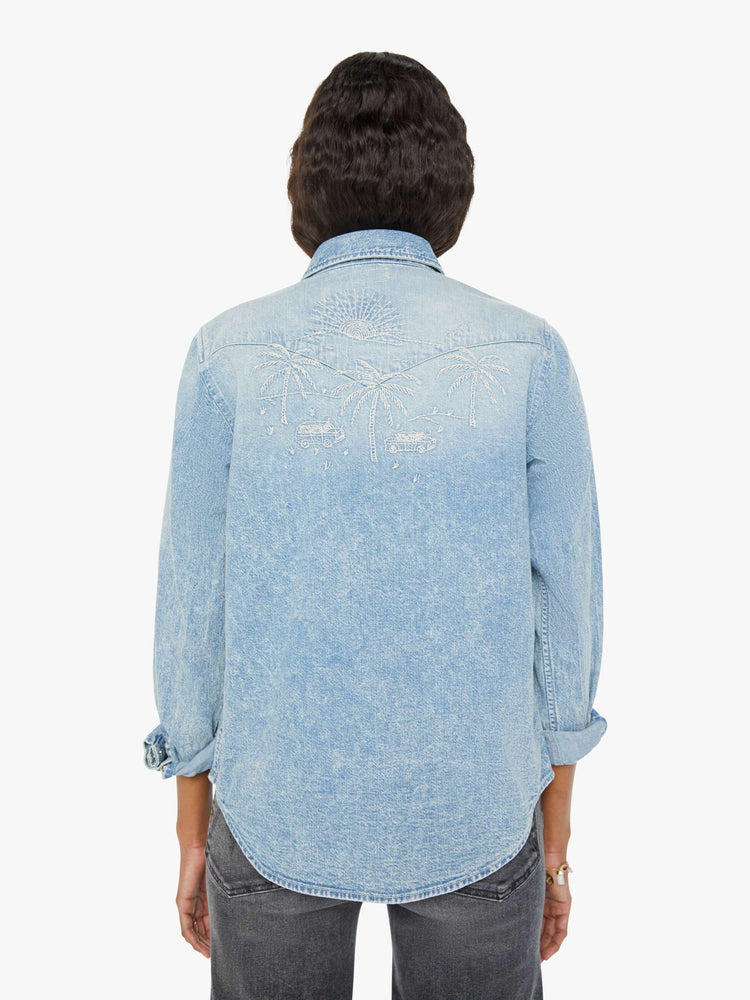 Back view of a woman in a light blue denim button-up with front patch pockets, a curved hem, and embroidered details throughout.