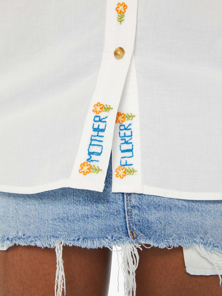 Detailed hem view of a woman in a white long sleeve button-up with colorful floral embroidery, as well as MOTHER and FUCKER toward the hem.