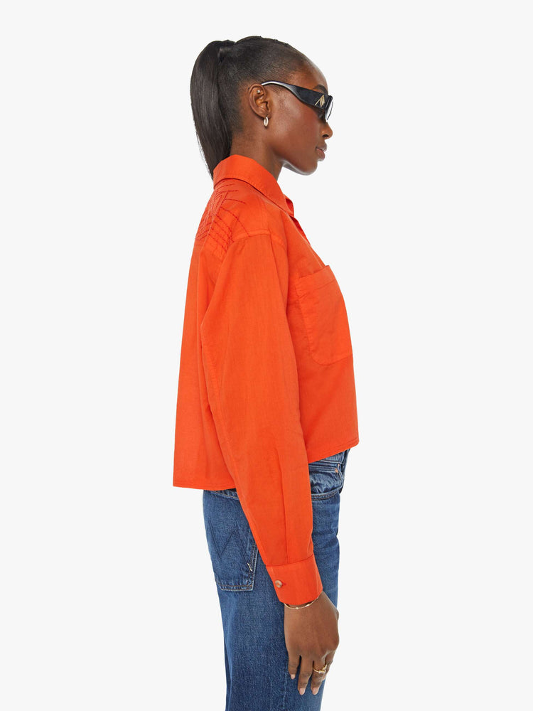 Side view of a woman in a long-sleeve bright orange collared button-up with drop shoulders, oversized patch pockets, a cropped, unfinished hem and MOTHER's name on the chest and mountains embroidered across the shoulders.