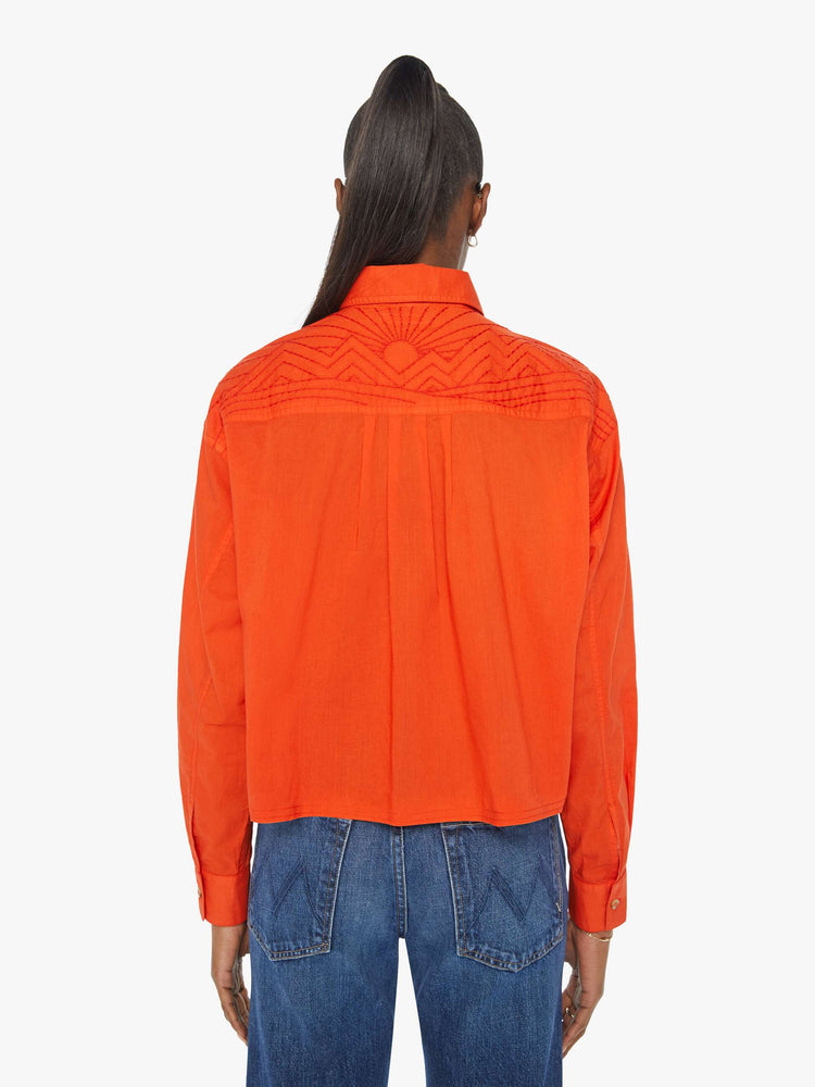 Back view of a woman in a long-sleeve bright orange collared button-up with drop shoulders, oversized patch pockets, a cropped, unfinished hem and MOTHER's name on the chest and mountains embroidered across the shoulders.