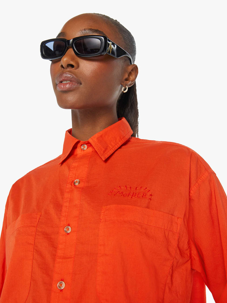 Front detailed view of a woman in a long-sleeve bright orange collared button-up with drop shoulders, oversized patch pockets, a cropped, unfinished hem and MOTHER's name on the chest and mountains embroidered across the shoulders.