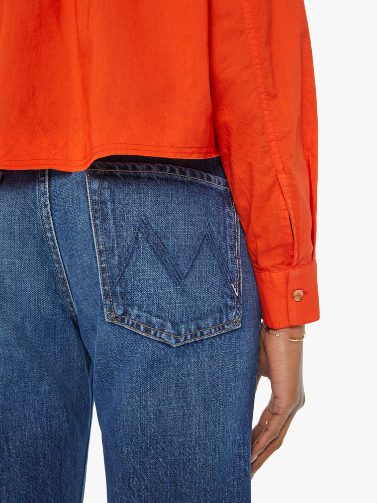 Detailed view of a woman in a long-sleeve bright orange collared button-up with drop shoulders, oversized patch pockets, a cropped, unfinished hem and MOTHER's name on the chest and mountains embroidered across the shoulders.