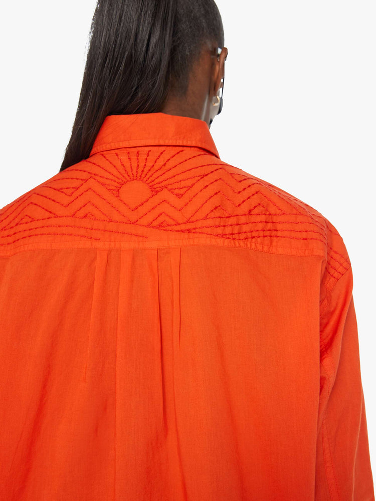 Back detailed view of a woman in a long-sleeve bright orange collared button-up with drop shoulders, oversized patch pockets, a cropped, unfinished hem and MOTHER's name on the chest and mountains embroidered across the shoulders.