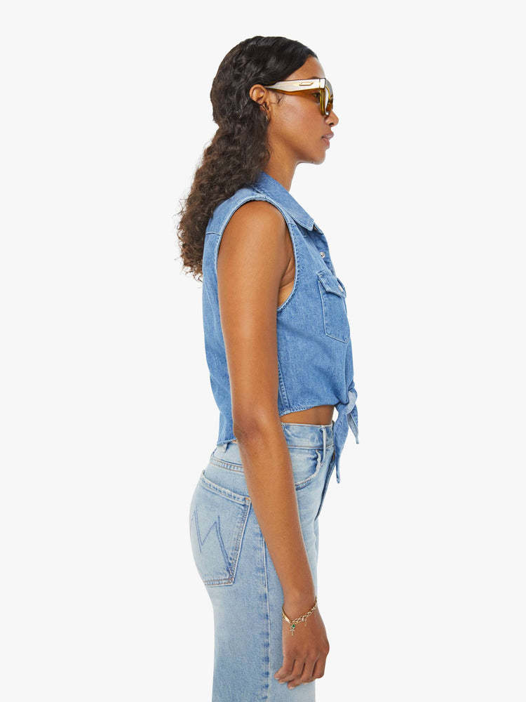 Side view of a woman medium blue sleeveless button-up with front patch pockets and a cropped hem that ties.