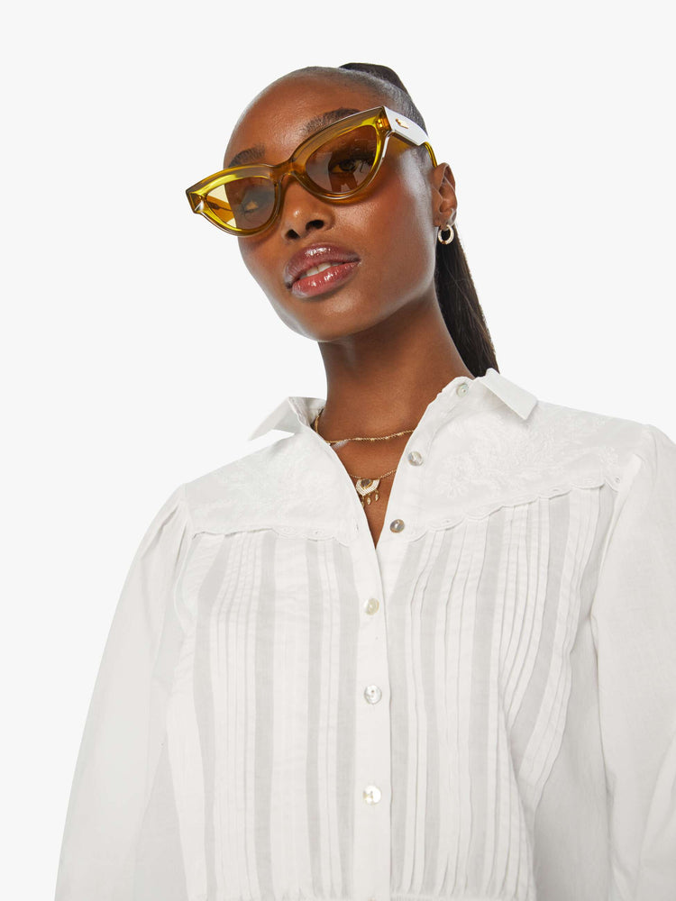 Close up view of a woman in a white blouse with puffed shoulders, elbow-length balloon sleeves, buttons down the front, and embroidered details throughout.