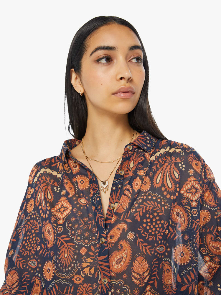 Close up view of a woman navy and orange paisley print button down blouse with a collared V-neck, 3/4 length balloon sleeves and curved hem.