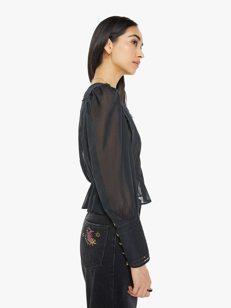 Side view of a woman in black blouse with long puff sleeves, a gathered elastic waist, long hems at the wrists that button and a cropped fit.