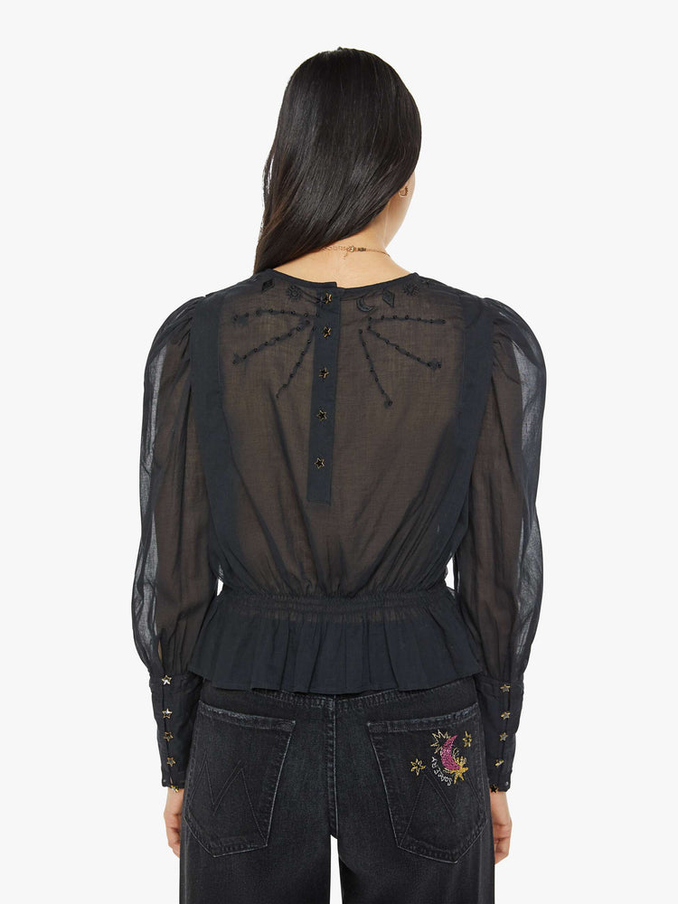 Back view of a woman in black blouse with long puff sleeves, a gathered elastic waist, long hems at the wrists that button and a cropped fit.