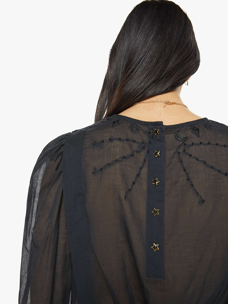 Back detailed view of a woman in black blouse with long puff sleeves, a gathered elastic waist, long hems at the wrists that button and a cropped fit.