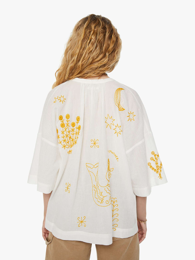 Back view of a woman in a white V-neck tunic with drop shoulders, elbow-length sleeves, front patch pockets and an oversized fit with yellow embroidered flowers, trees and animals throughout.