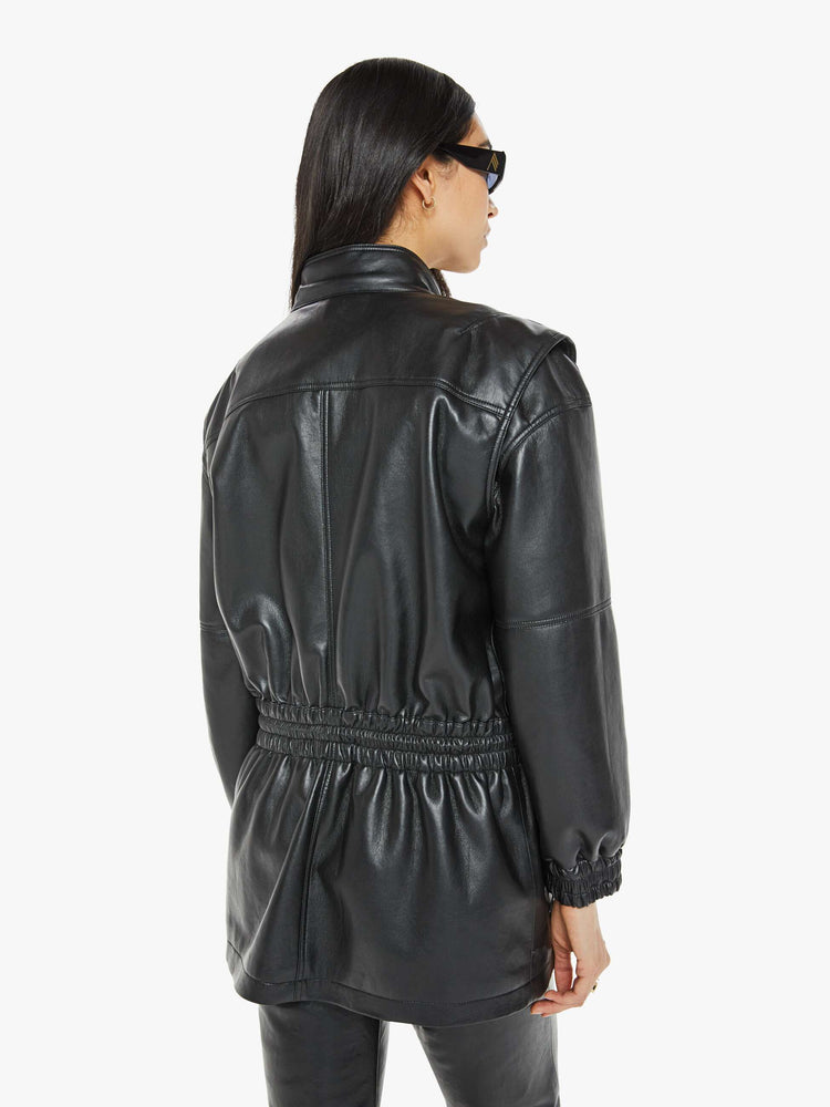 Back view of a woman in a leather black parka  with a stacked collar, patch pockets, elastic at the waist and wrists, a zip closure and drawstring hem that hits mid-thigh. 