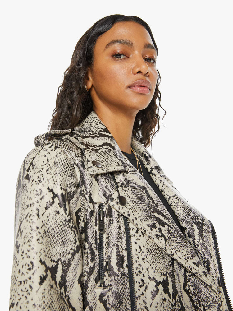 Close up view of a woman classic motorcycle jacket with a notched collar, zip pockets, belted waistband and a slightly cropped fit in a cream and black snake print.