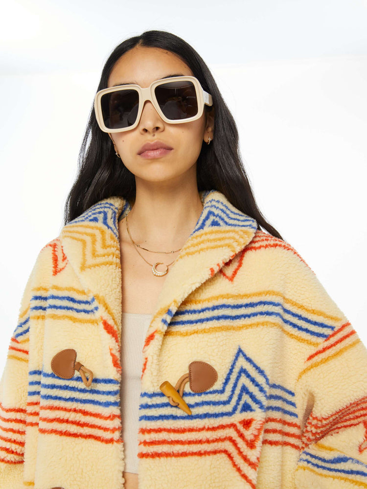 Detailed view of a woman in a light tan faux sherpa oversized jacket with a horizontal stripe pattern in red, blue and yellow, patch pockets, three horn buttons down the front.