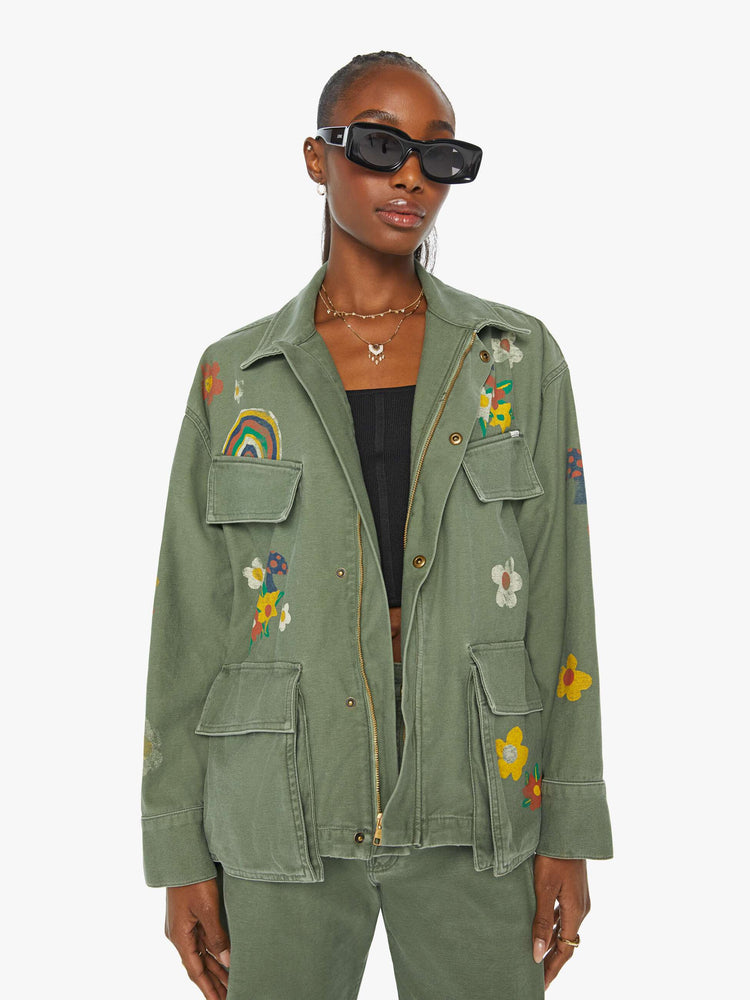 Front view of a woman in a military green flower and rainbow embroidered collared jacket with oversized patch pockets, a zip closure that snaps shut.