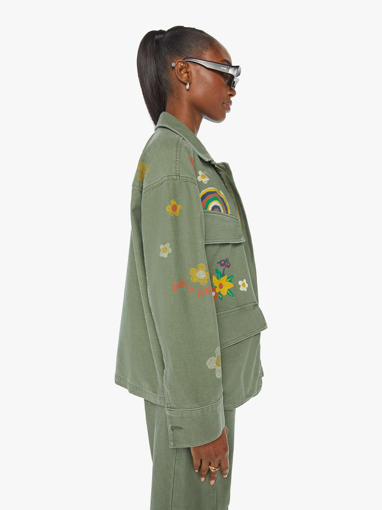 Side view of a woman in a military green flower and rainbow embroidered collared jacket with oversized patch pockets, a zip closure that snaps shut.