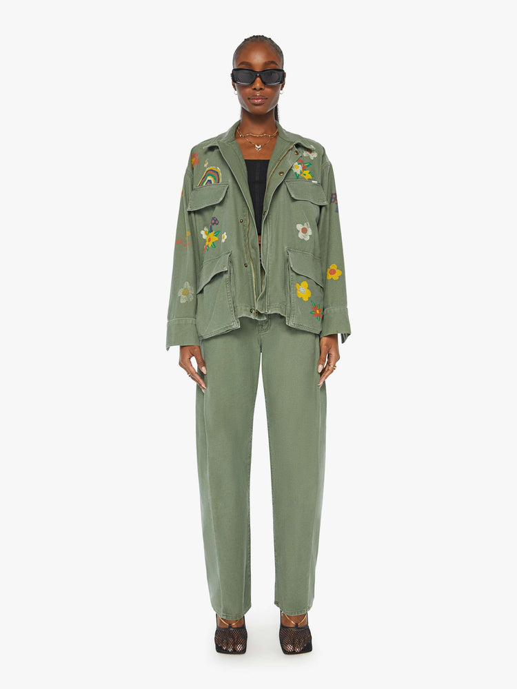 Full front view of a woman in a military green flower and rainbow embroidered collared jacket with oversized patch pockets, a zip closure that snaps shut.