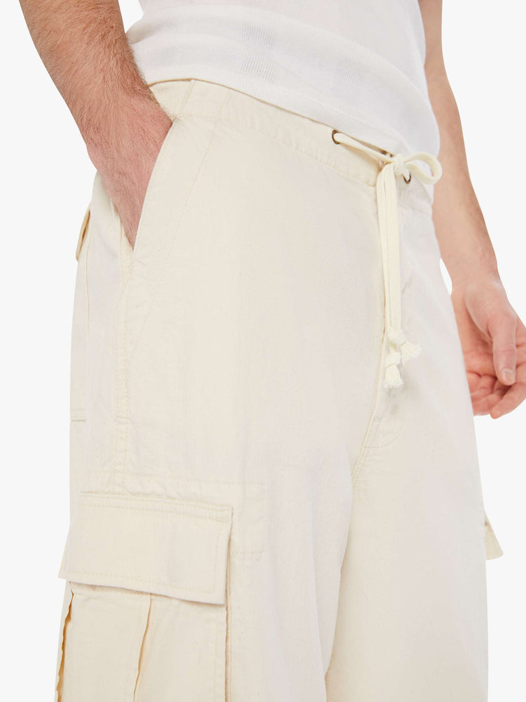 Close up view of a men in a creamy white drawstring cargo pants with a loose wide-leg, patch pockets, a 26.5-inch inseam and ties at the hems.