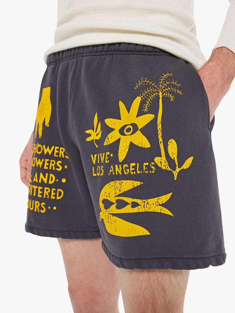 Close up view of a man's faded black with yellow graphic sweatshorts with a loose wide leg, 7.5-inch inseam and a raw hem.