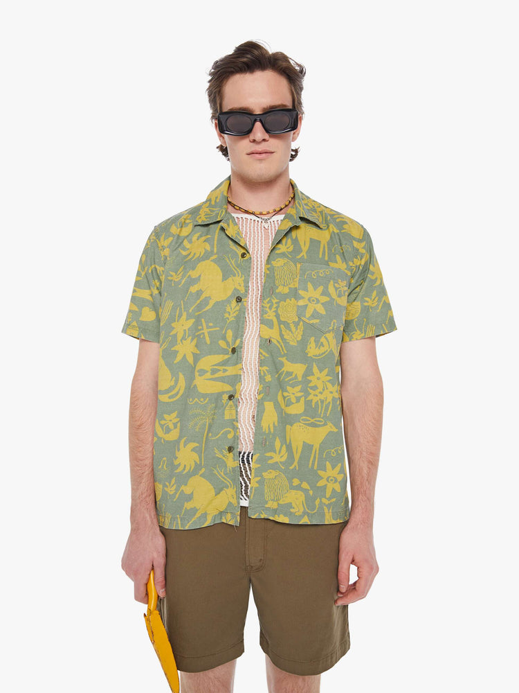 Front view of a man in a sage green short sleeve button-up with a patch pocket and notched collar designed with printed animals, flowers and symbols in yellow.