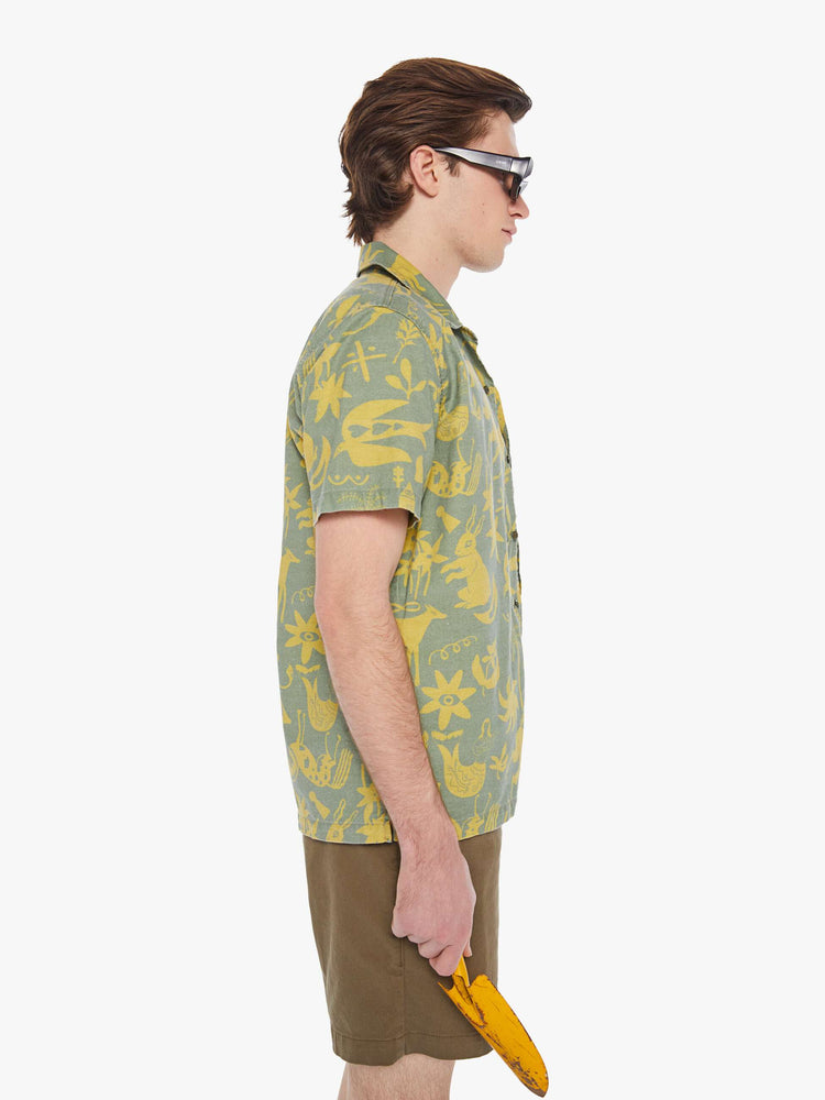 Side view of a man in a sage green short sleeve button-up with a patch pocket and notched collar designed with printed animals, flowers and symbols in yellow.