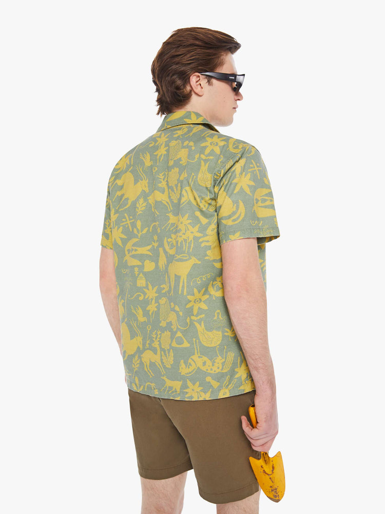 Back view of a man in a sage green short sleeve button-up with a patch pocket and notched collar designed with printed animals, flowers and symbols in yellow.