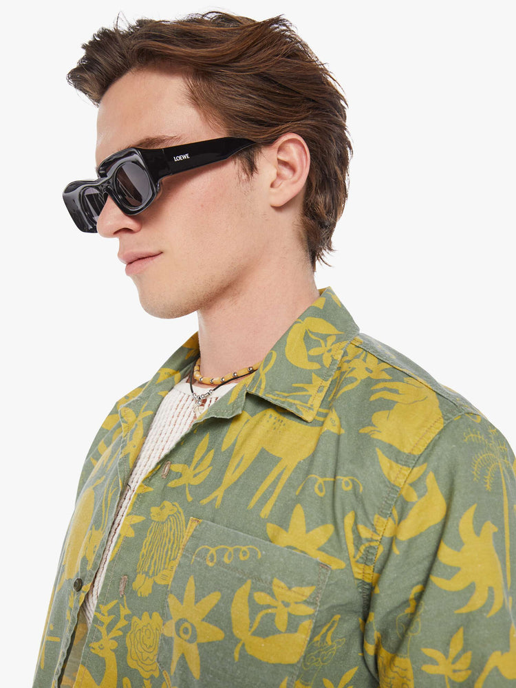 Detailed view of a man in a sage green short sleeve button-up with a patch pocket and notched collar designed with printed animals, flowers and symbols in yellow.